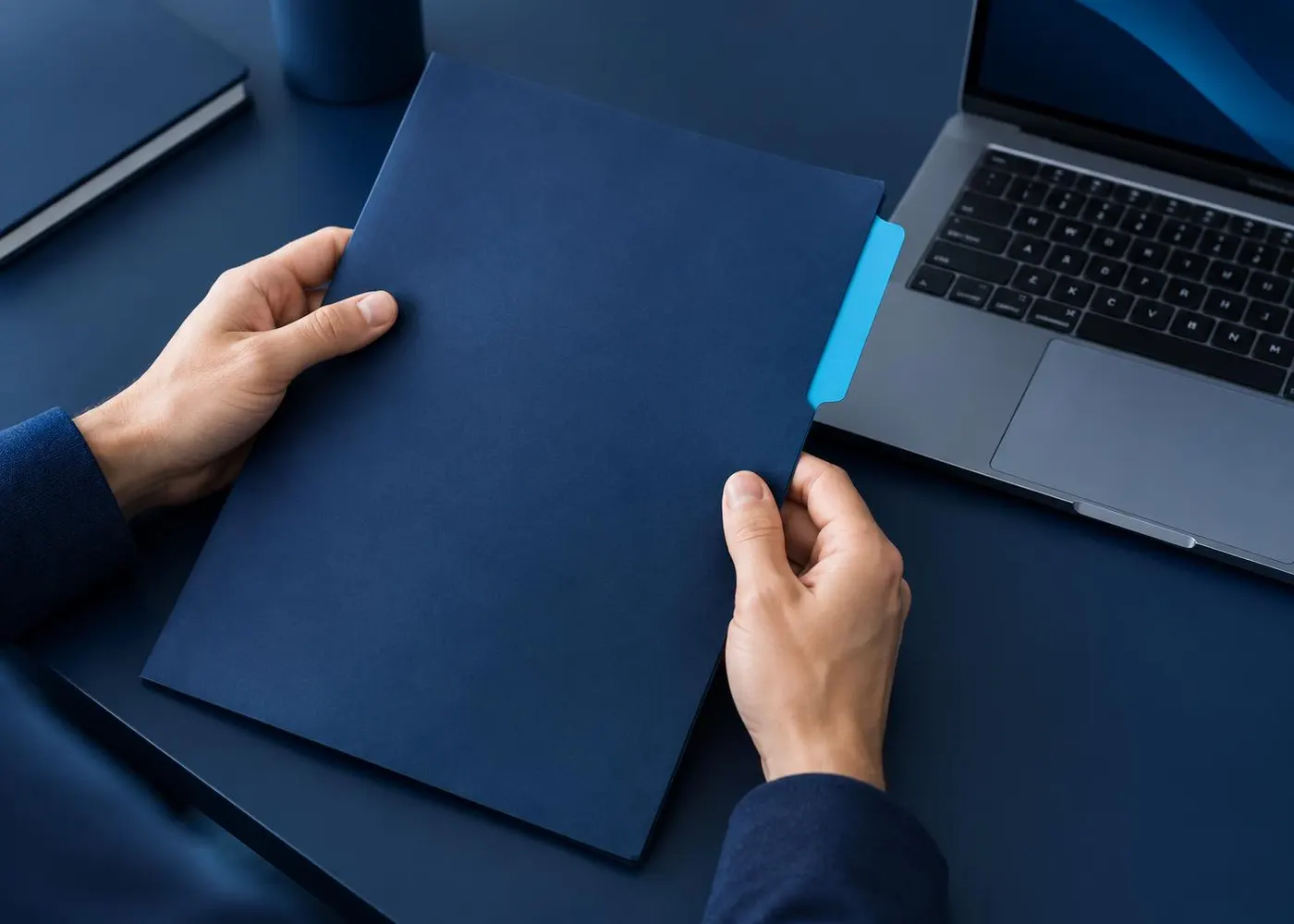 Hands holding a navy folder with a cyan tab next to a laptop